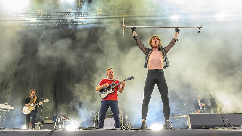 A live performance from Cage the Elephant in 2019 with the lead singer holding up the mic stand above his head.