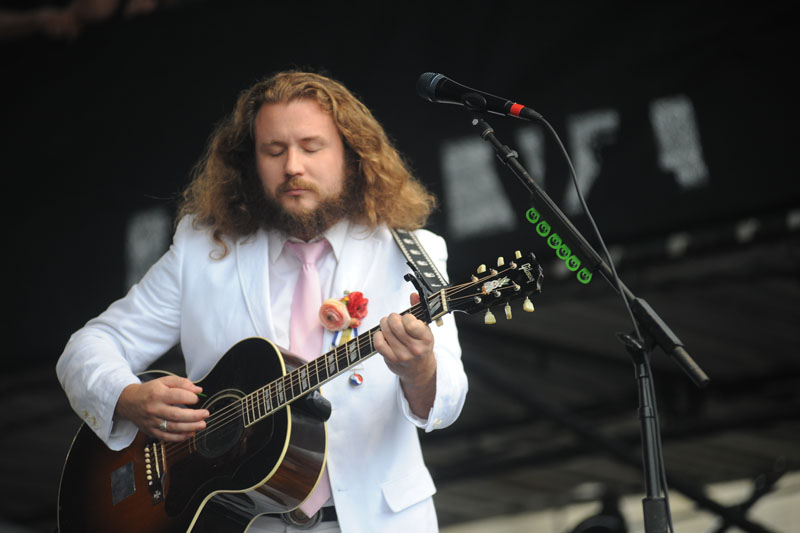Jim James from My Morning Jacket wearing a white suite with a pink tie playing a dark-colored acoustic guitar