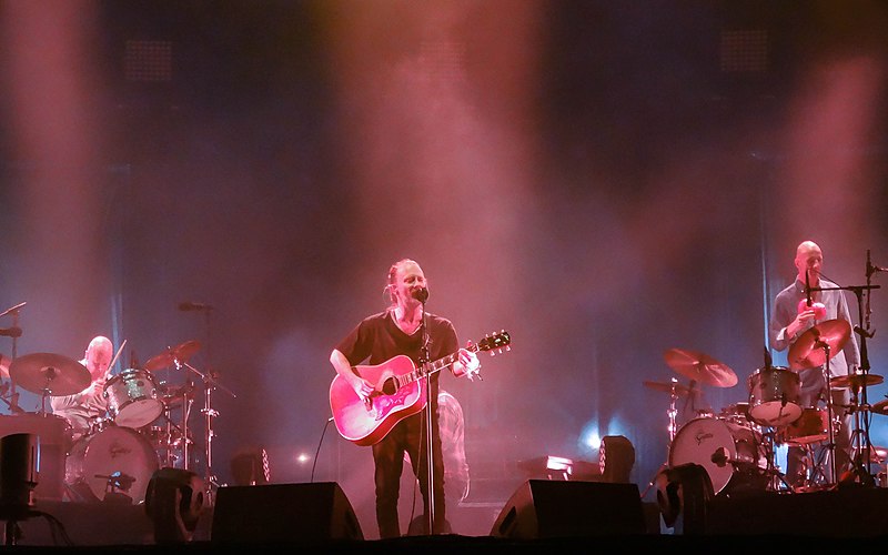 Thom Yorke from Radiohead playing an acoustic guitar and singing on stage with Radiohead drummer Phil Selway and additional percussionist.